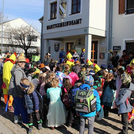 Eine Gruppe von Kindern in Kostümen und Hüten versammelt sich vor einem Gebäude. Einige Erwachsene in Kostümen stehen auf den Stufen, einer hält ein Musikinstrument. Das Gebäude hat Schilder und ein Wappen.
