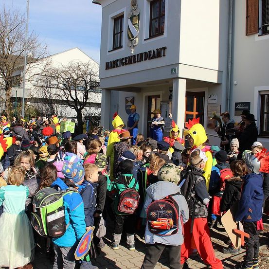 Eine Menschenmenge von Kindern und Erwachsenen in Kostümen und Mützen versammelt sich vor einem Gebäude. Einige tragen Hühnerkostüme, während andere Mützen und Rucksäcke haben. Einige Personen spielen Instrumente.