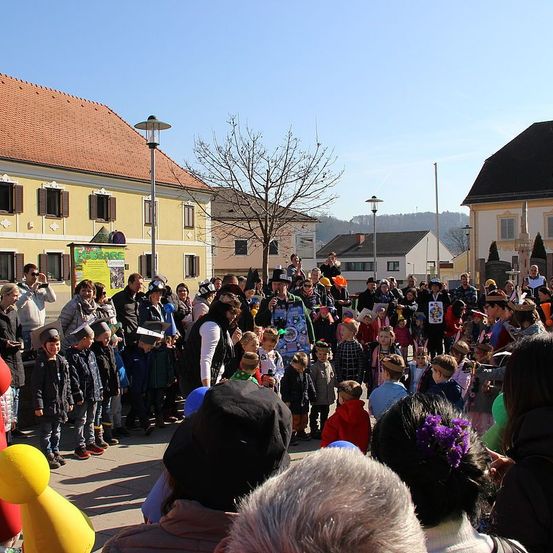 Eine Gruppe von Menschen versammelt sich an einem Ort im Freien, wahrscheinlich einem Stadtplatz, mit Gebäuden im Hintergrund. Einige tragen Hüte, und einige haben Luftballons. Ein Mann in Kostüm steht vorne und spricht die Menge an.
