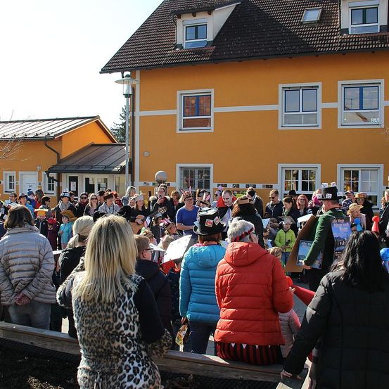 Eine Menschenmenge, einige mit Hüten, versammelt sich vor einem gelben Gebäude. Ein Mann in einem schwarzen Hut spielt Gitarre, während andere zusehen. Einige Leute sitzen auf einer Bank, und eine Straßenlampe ist in der Nähe.