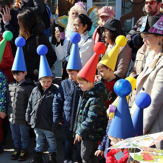 Eine Gruppe von Kindern mit blauen, roten und gelben Kegelmützen steht in einer Reihe, mit Erwachsenen und anderen Kindern um sie herum. Ein Erwachsener macht ein Foto mit einem Handy.