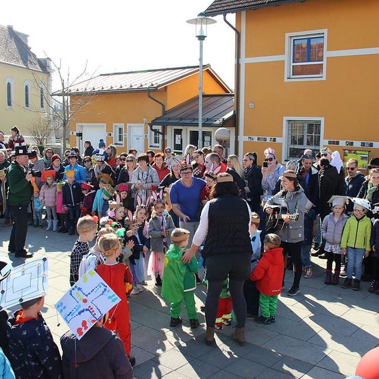 Eine Menschenmenge, darunter Kinder in Kostümen, versammelt sich im Freien. Eine Person in der Mitte spielt Gitarre, während andere zusehen. Hinter ihnen befinden sich einige Gebäude mit Glasfenstern.
