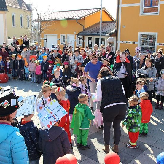 Eine Menschenmenge, bestehend aus Kindern und Erwachsenen, versammelt sich vor Gebäuden, einige in Kostümen und Hüten. Eine Person spielt Gitarre.