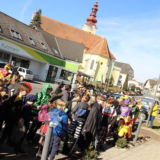 Viele Kinder in Kostümen versammeln sich vor einer Kirche und einem Geschäft unter klarem Himmel. Gebäude und geparkte Autos sind im Hintergrund zu sehen.