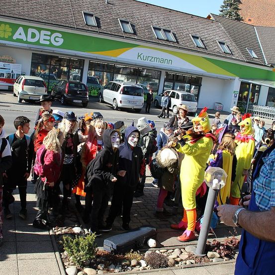 Eine Gruppe von Menschen in verschiedenen Kostümen versammelt sich vor einem Kurzmann-Geschäft mit geparkten Autos. Einige tragen Masken und Hüte, während andere Instrumente spielen.