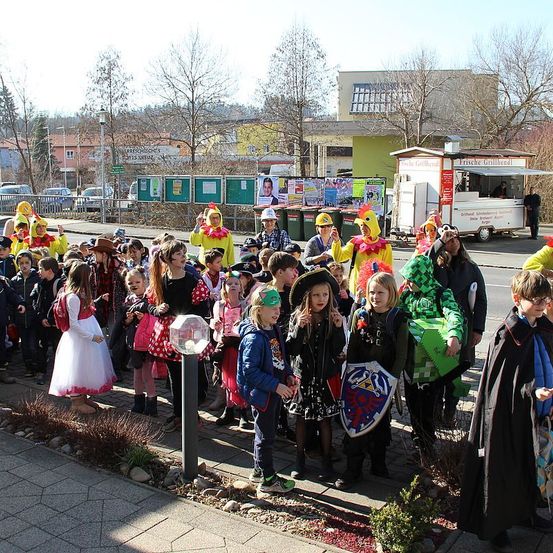 Eine Gruppe von Kindern in Kostümen steht auf einem Bürgersteig, einige tragen Hüte und halten Schilde. Im Hintergrund befinden sich Gebäude, Autos und ein Imbisswagen.