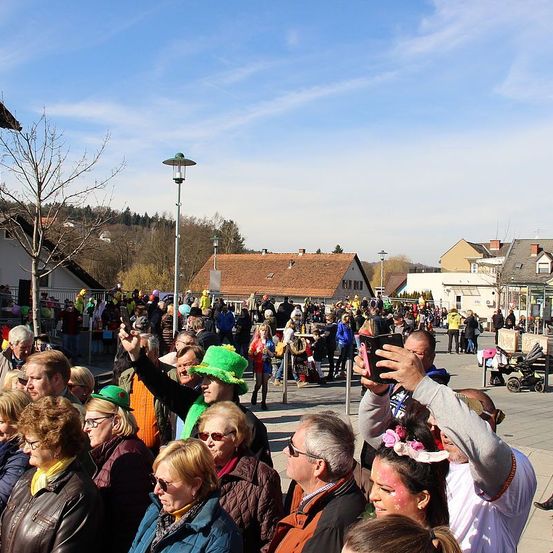 Eine große Menschenmenge auf einem Stadtplatz, einige in Kostümen gekleidet, an einem sonnigen Tag. Ein Mann macht ein Foto mit seinem Handy.
