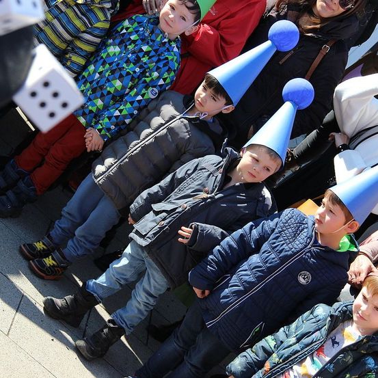 Eine Gruppe von Kindern in Winterkleidung, einige mit blauen Partyhüten, sitzen eng zusammen auf einem gepflasterten Bereich, möglicherweise bei einer öffentlichen Veranstaltung.