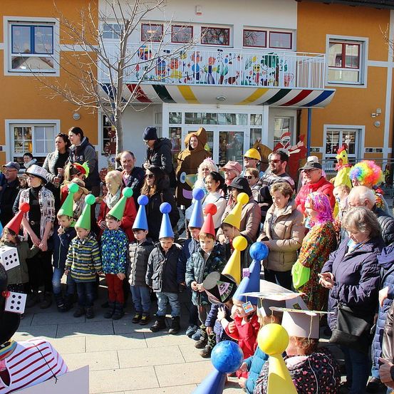 Eine Menschenmenge, bestehend aus Kindern und Erwachsenen, versammelt sich vor einem Gebäude mit bunten Dekorationen, hält Luftballons und trägt Hüte. Die Kinder tragen bunte Hüte, während einige Erwachsene in Kostümen sind. Das Gebäude hat einen Balkon mit einem farbenfrohen Wandbild.