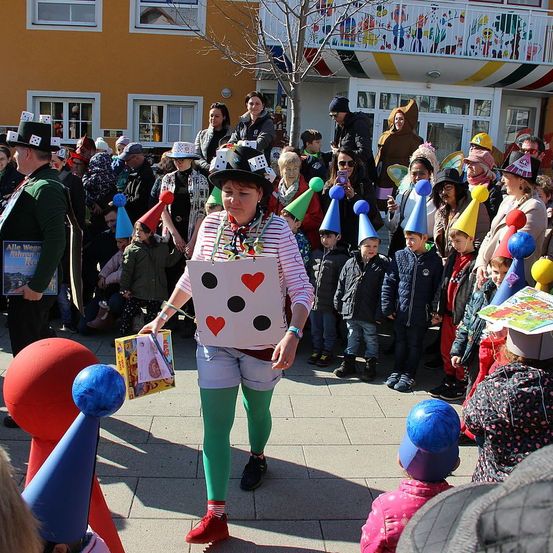 Eine Gruppe von Menschen, einige in Kostümen, versammelt sich im Freien. Eine Frau in einer Spielkarten-Kostüm hält eine Schachtel. Kinder in Partyhüten stehen davor. Dahinter schauen andere zu, und Gebäude mit bunten Dekorationen sind sichtbar.