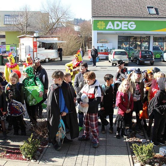 Eine Gruppe von Kindern in Kostümen versammelt sich auf einem Bürgersteig vor einem Geschäft, mit geparkten Autos in der Nähe und einem mobilen Imbisswagen im Hintergrund.