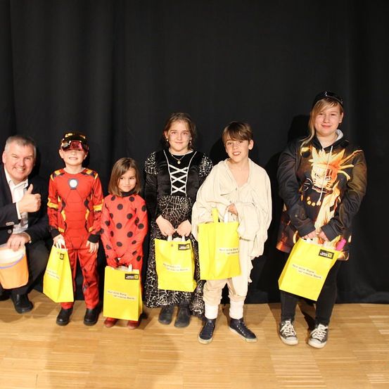 Eine Gruppe von Kindern und einem Erwachsenen posiert für ein Foto, gekleidet in verschiedenen Kostümen, mit gelben Taschen in der Hand. Sie stehen auf einem Holzboden vor einem schwarzen Hintergrund.