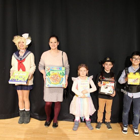 Fünf Kinder in verschiedenen Kostümen stehen auf einer Bühne und halten verschiedene Spielzeuge in den Händen. Eine Frau in der Mitte hält ein Spielzeug mit 'Slime XXL' darauf. Die Bühne hat einen Holzboden und einen schwarzen Hintergrund.