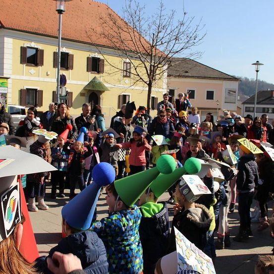 Eine Menschenmenge, darunter Kinder mit Partyhüten, versammelt sich im Freien. Einige tragen Jacken und Stiefel, andere halten Schilder. Im Hintergrund steht ein Gebäude mit mehreren Fenstern und einem Baum.