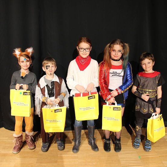 Fünf Kinder in verschiedenen Kostümen posieren für ein Foto. Jedes hält einen gelben Beutel, von zweien ist der Text sichtbar. Sie stehen vor einem schwarzen Hintergrund.