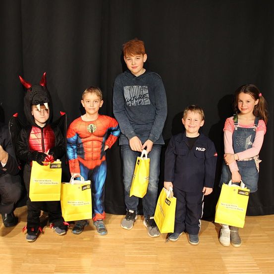 Eine Gruppe von Kindern in Kostümen, die gelbe Taschen halten und für ein Foto auf einem Holzboden posieren. Ein Junge in einem Spider-Man-Kostüm steht zwischen einem Jungen in einem Teufelskostüm und einem Mädchen in einer Latzhose.