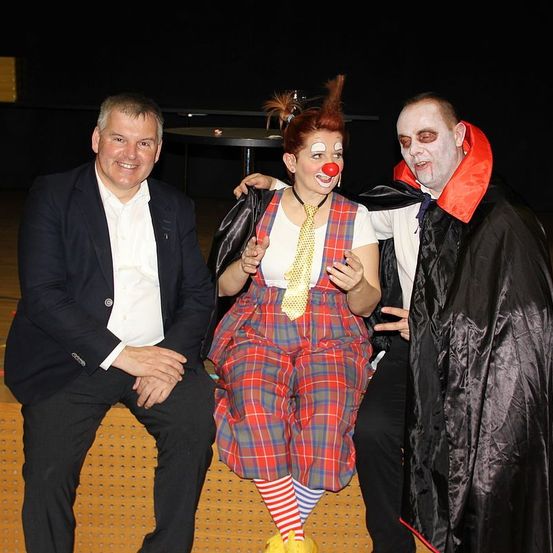 Drei Menschen in Kostümen. Einer ist ein Clown, einer ein Vampir und einer ein Mann in Anzug, der lächelt.