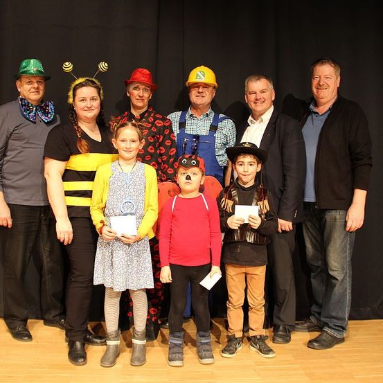 Eine Gruppe von Erwachsenen und Kindern posiert für ein Foto auf der Bühne. Sie tragen Kostüme, manche halten Urkunden. Der Hintergrund ist ein schwarzer Vorhang.
