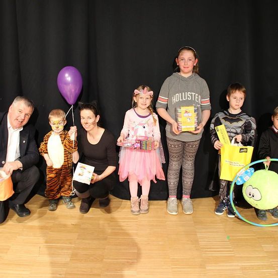Eine Gruppe von Kindern und Erwachsenen posiert für ein Foto auf einer Bühne, alle halten Geschenke. Ein Mann in Anzug kniet auf der linken Seite, während ein Mädchen in einer rosa Tutu und ein Junge in einer Batman-Kostüm in der Mitte stehen.