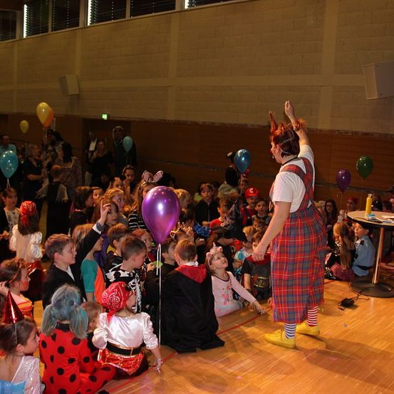 Ein Saal voller Kinder, einige in Kostümen, beobachten einen Clown mit einem lila Luftballon auf der Bühne. Hinter ihnen stehen Menschen und halten Luftballons. Der Raum hat Ziegelwände und Glasfenster.