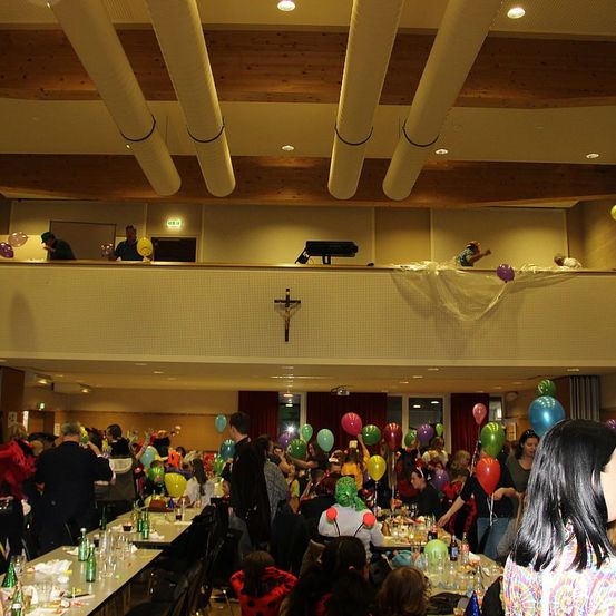 Eine große Indoor-Veranstaltung mit mehreren Personen, Tischen und Luftballons. Einige Personen tragen Kostüme und stehen auf einer Balkonseite.