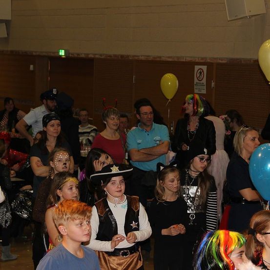 Eine Gruppe von Menschen ist drinnen versammelt, wahrscheinlich auf einer Kostümparty. Einige tragen Gesichtsbemalung und Kostüme. Eine Frau mit farbenfrohen Haaren und einem Piratenhut sticht hervor. Luftballons und ein Schild sind sichtbar.