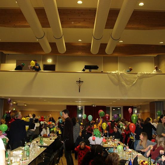Ein Indoor-Veranstaltungsort ist voller Teilnehmer. Viele Menschen sind in Kostümen gekleidet und halten bunte Luftballons. Tische sind mit Getränken und Essen beladen. Der Raum verfügt über Holzbalken an der Decke und eine Galerie.