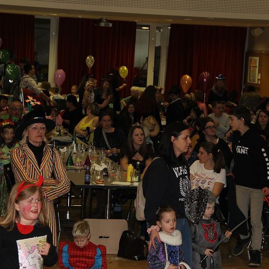 Ein überfüllter Raum mit Luftballons und Menschen in Kostümen, einige mit Kindern, die Schwerter und Schilde halten. Eine Frau in einer Hexenkostüm und ein Mann in einem schwarzen Shirt. Es gibt Tische mit Flaschen, Gläsern und anderen Gegenständen.