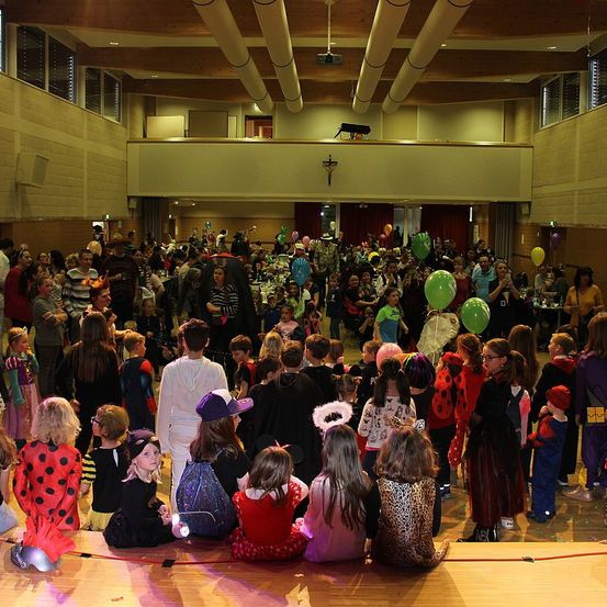 Eine große Gruppe von Kindern und Erwachsenen ist in einem Saal für eine Halloween-Veranstaltung versammelt. Viele sind in Kostümen gekleidet, und es gibt Ballons und Dekorationen in der Luft.