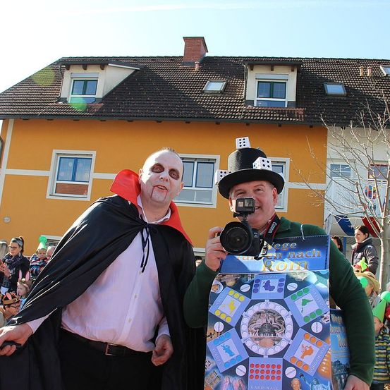 Zwei Männer in Kostümen stehen vor einem Gebäude mit Publikum. Ein Mann ist als Dracula verkleidet und der andere trägt einen Hut mit Würfeln. Der Mann im Dracula-Kostüm hält eine Kamera.