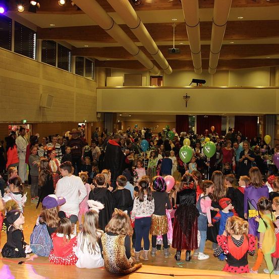 Eine große Gruppe von Kindern in Kostümen versammelt sich in einem Saal, einige sitzen und andere stehen, mit bunten Luftballons und einer Holzdecke.