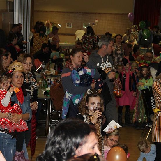 Eine große Menschenmenge in einem Saal, viele in Kostümen, halten Getränke und Ballons. Einige halten Kinder, und einige tragen Brillen. Tische sind mit Flaschen und Essen besetzt. Der Raum hat Ballons und Dekorationen.