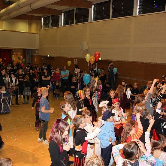 Eine Gruppe von Kindern und Erwachsenen versammelt sich in einem Raum, wahrscheinlich für eine Party. Einige von ihnen tragen Kostüme und einige halten Luftballons. Der Raum ist mit bunten Konfetti auf dem Boden dekoriert. Es gibt auch Menschen im Hintergrund, die die Kinder beobachten.
