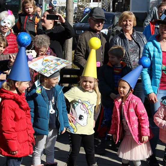 Eine Gruppe von Kindern mit Partyhüten, die für die Kamera lächeln. Dahinter machen Erwachsene Fotos mit ihren Handys. Eine Frau in einer Maske spielt Gitarre.