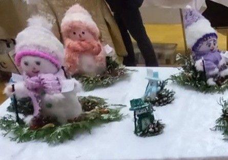 Ein Tisch mit einem weißen Tuch ist mit mehreren handgefertigten Schneemanndekorationen bedeckt. Jeder Schneemann ist einzigartig und mit verschiedenen Schals, Mützen und Accessoires geschmückt. Einer hält eine Laterne, während andere kleine Kiefernzapfen und Zweige haben. Eine Person im Hintergrund trägt schwarze Hosen.
