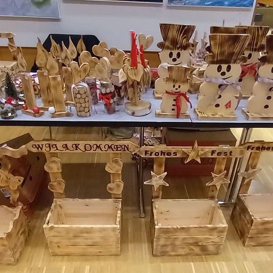 Ein Holztisch zeigt verschiedene handgefertigte Gegenstände, darunter Schneemänner, Holzkisten und Sterne. Es gibt ein Schild mit der Aufschrift 'WILLKOMMEN' und ein weiteres mit der Aufschrift 'Frohes Fest'. Der Tisch steht auf einem Holzboden.