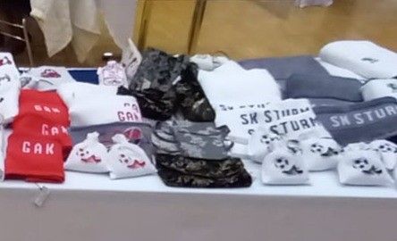 Ein Tisch zeigt verschiedene Artikel, darunter Socken, T-Shirts und Taschen. Einige Artikel haben rote, weiße und graue Farben. Ein Artikel hat das Wort 'GAK' und ein anderer 'SK STURM'.