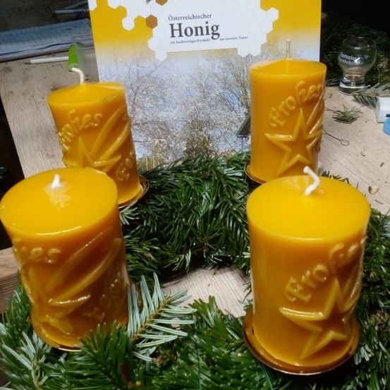 Vier gelbe Kerzen sind in einem Kreis mit einem Kranz angeordnet, wobei eine Kerze mit dem Wort 'Euro' graviert ist. Ein Buch ist hinter den Kerzen platziert. Ein Glas steht rechts. Ein Stift und ein Stück Papier liegen auf dem Tisch.