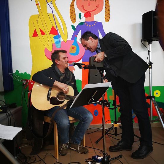 Zwei Männer sind drinnen, einer spielt Gitarre und der andere lacht. Sie stehen vor einer bunten Wand mit einer Illustration und einem Mikrofon.