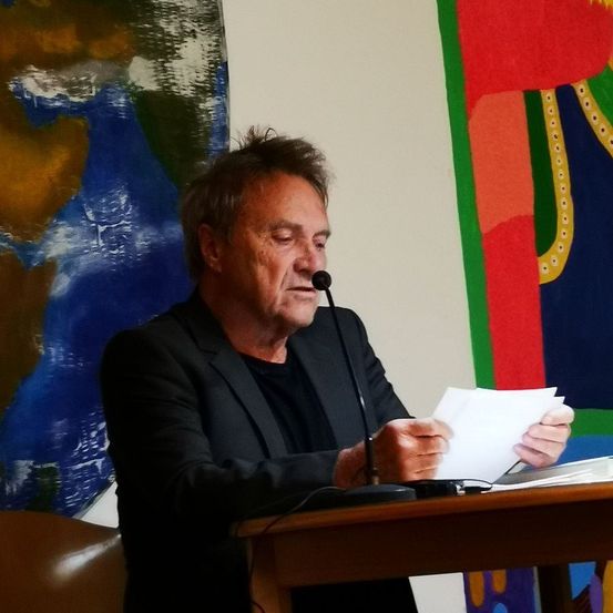 Ein Mann in einem schwarzen Anzug sitzt an einem Podium, hält Papiere in der Hand und spricht in ein Mikrofon. Hinter ihm hängen zwei große Gemälde an der Wand.