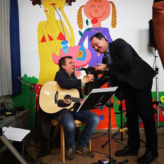 Zwei Männer spielen Musik in einem Raum mit einem farbenfrohen Wandbild. Ein Mann spielt Gitarre, während der andere ein Mikrofon hält. Sie scheinen beide viel Spaß zu haben.