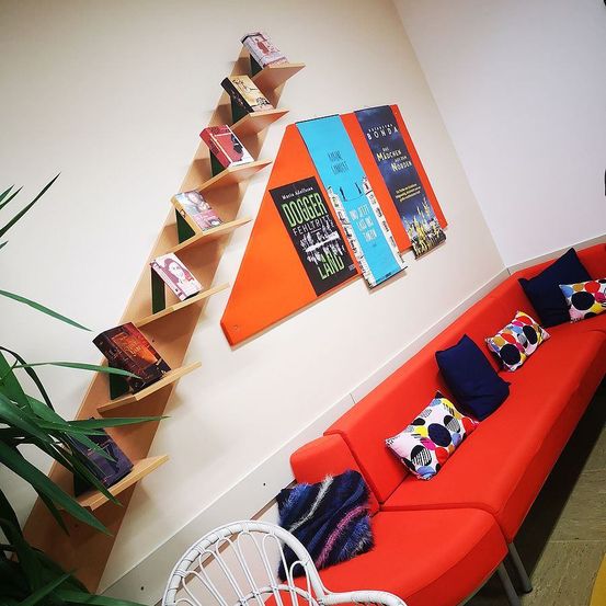 Ein gemütliches Zimmer mit einem roten Sofa, dekorativen Kissen und einem dreieckigen Bücherregal mit Büchern. Die Wand ist mit einem weißen Hintergrund, drei bunten Postern und einer grünen Pflanze auf der linken Seite geschmückt.