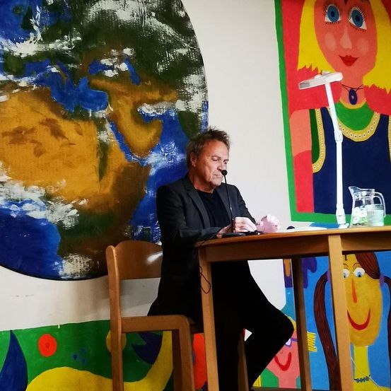 Ein Mann in einem schwarzen Anzug sitzt an einem Schreibtisch und schaut auf ein Mikrofon. Hinter ihm ist eine Weltkugel an der Wand gemalt, und rechts ist das Gesicht einer Frau.