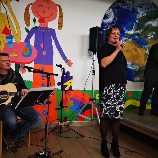 Eine Frau singt in ein Mikrofon auf der Bühne. Ein Mann spielt Gitarre neben ihr. Bunte Wandgemälde hängen an der Wand hinter ihnen.