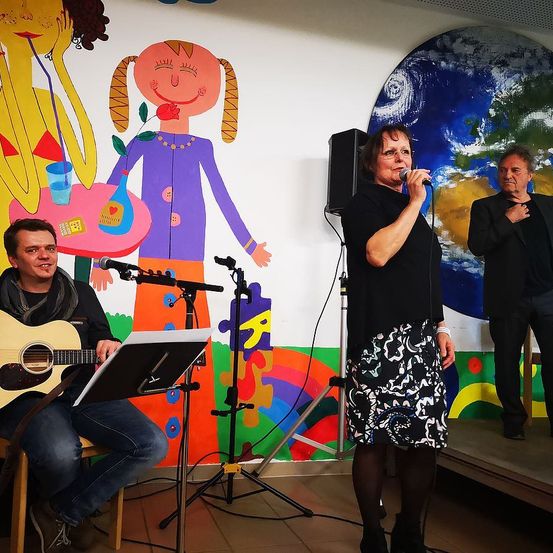 Drei Personen auf der Bühne mit Musikinstrumenten. Eine Frau singt in ein Mikrofon. Ein Mann spielt die Gitarre. Ein anderer Mann steht hinter ihr. Eine farbenfrohe Wandmalerei befindet sich an der Wand hinter ihnen.
