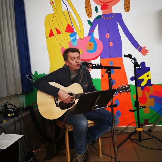 Ein Mann sitzt auf einem Stuhl und spielt eine Gitarre, während er in ein Mikrofon singt vor einer farbenfrohen Wandmalerei.