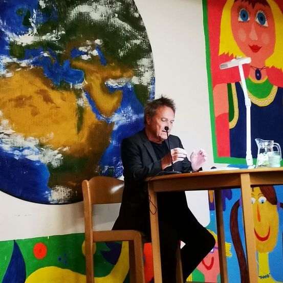 Ein Mann in einem Anzug sitzt an einem Tisch mit einem Mikrofon und hält eine Rede mit einem Gemälde der Erde im Hintergrund.