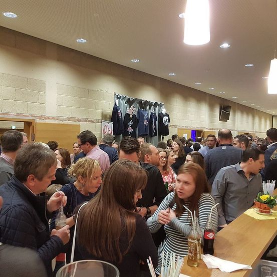 Ein überfüllter Innenbereich zeigt eine Bar mit Flaschen, Bechern und Strohhalmen. Mehrere Menschen sind versammelt, unterhalten sich und trinken. Es hängen T-Shirts an der Wand und ein Schild.