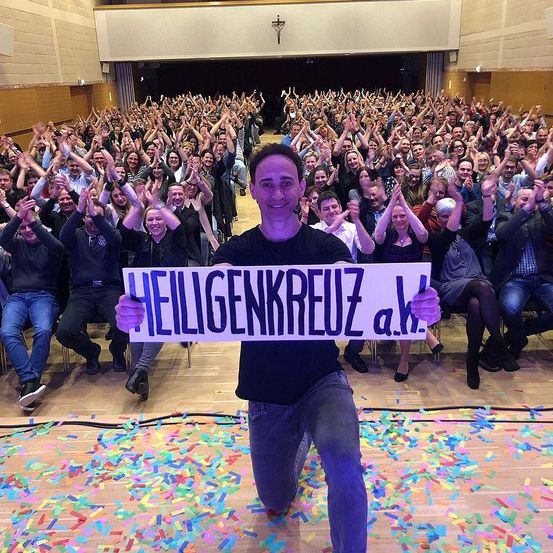 Ein Mann hält ein Schild mit der Aufschrift Heiligenkreuz a.k.a. in der Knie vor einer jubelnden Menge in einem großen Auditorium.