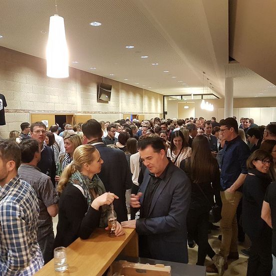 Eine Menschenmenge in einer Bar, einige halten Getränke, andere unterhalten sich, unter einer beleuchteten Decke und einer Wand mit Ziegeldesign.